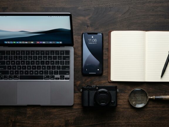 Notebook aberto, smartphone e câmera compacta sobre uma mesa de madeira com um caderno em branco.